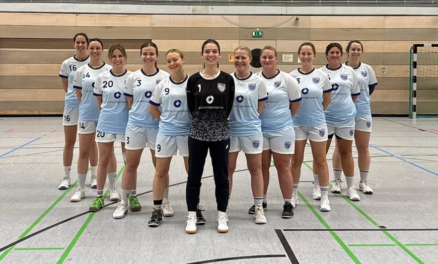 Eine Handballmannschaft in blauen und weißen Trikots steht in einer Halle, mit einer Spielerin in Torwartkleidung in der Mitte.