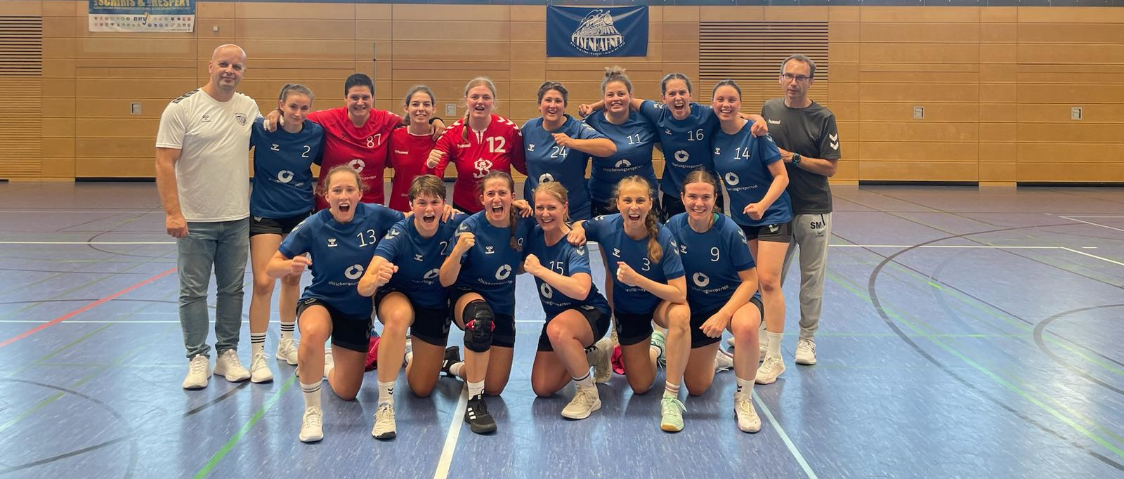 Ein Handballteam posiert lächelnd in einer Sporthalle.